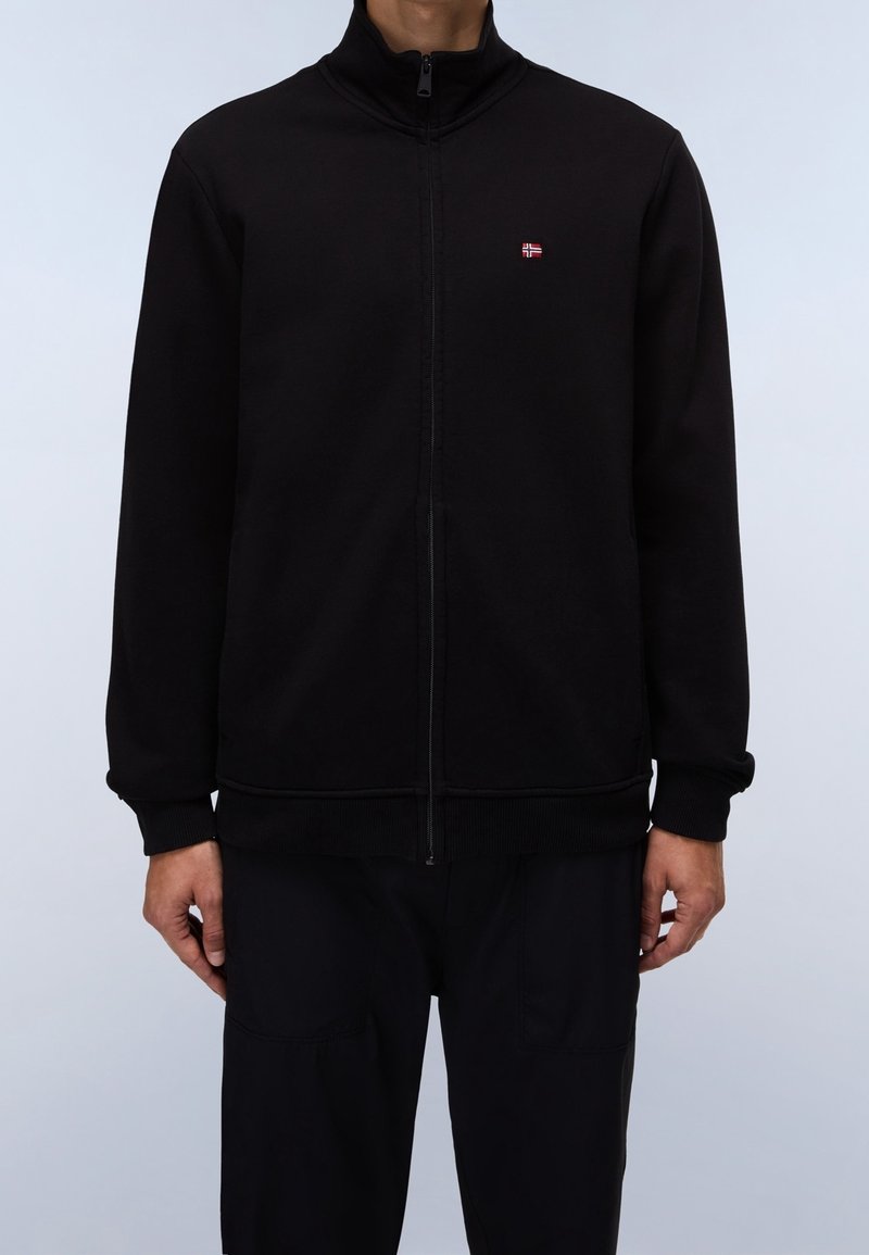Schwarze Zip-Jacke mit hohem Kragen, aus weichem Stoff gefertigt. Verfügt über ein kleines rotes Logo auf der linken Brust. Gerippte Bündchen und Saum.