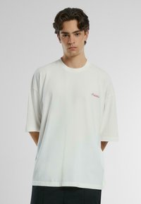 Camiseta de algodón blanca con mangas cortas, cuello redondo y "Prohibido" bordado en rojo en el lado izquierdo. Textura suave, corte relajado.