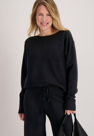 Zwarte gebreide trui met een brede halslijn, lange mouwen en geribde manchetten, gecombineerd met bijpassende zwarte broek. Model houdt een zwarte handtas vast.