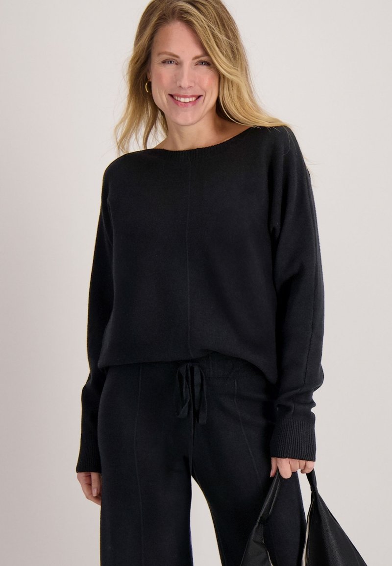 Zwarte gebreide trui met een brede halslijn, lange mouwen en geribde manchetten, gecombineerd met bijpassende zwarte broek. Model houdt een zwarte handtas vast.