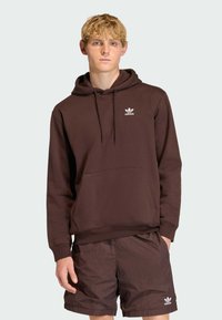 Hnedá mikina s kapucňou a bielym logom Adidas, s predným vreckom, šnúrkami a ribbed manžetami, v kombinácii s hnedými šortkami.