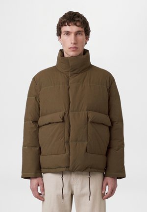 Un homme porte une veste matelassée brun olive, à col montant et grandes poches avant, sur un fond blanc uni.