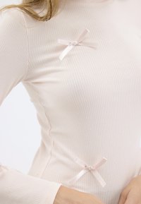Tynn ribbestrikket topp i myk rosa, med to små satengsløyfer i matchende farge foran. Lange ermer, tettsittende design, glatt tekstur.