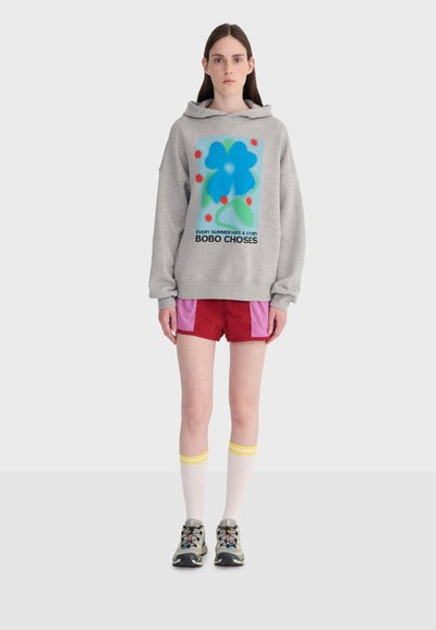 Jeune femme portant un sweat à capuche gris avec une fleur bleue et un texte, un short rouge et rose, des chaussettes blanches montantes avec des rayures jaunes, et des baskets.