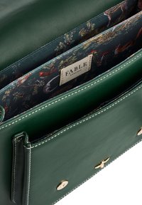 Borsa in pelle verde con accessori dorati, cuciture bianche e fodera in tessuto con motivo che presenta animali e natura. Etichetta del marchio visibile.