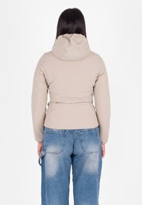 Vrouw met lang bruin haar, gekleed in een beige hoodie en vervaagde blauwe denim jeans, staand met haar rug naar de camera tegen een witte achtergrond.