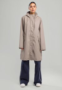 Vedennepitävä beige sadetakki, jossa on huppu, edessä napituskotelo ja sivutaskut. Päällä siniset leveälahkeiset housut ja valkoiset lenkkarit.