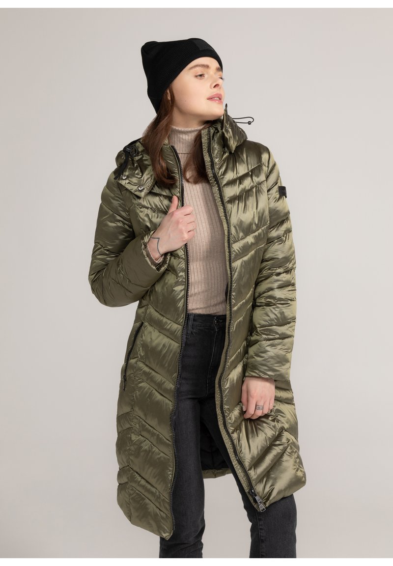Lange olijfgroene pufferjas met een doorstikt ontwerp, gewatteerde zakken en een hoge kraag. Draagt over een beige coltrui en zwarte broek.