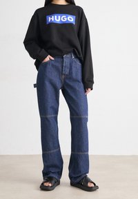 Zwarte sweatshirt met een blauw "HUGO" logo, gecombineerd met blauwe spijkerbroeken met wijde pijpen en beige stiksel. Zwarte sandalen maken de look compleet.