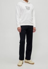 Felpa bianca con cappuccio e logo "Emporio Armani", abbinata a pantaloni blu navy e sneakers beige; tessuto liscio, vestibilità rilassata, design minimalista.