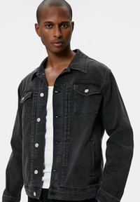 Zwart denim jacket met een knoopsluiting aan de voorkant, lange mouwen, twee borstzakken en een klassieke kraag. Subtiele stikdetails accentueren het ontwerp.
