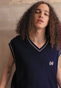 Navy sleeveless knit top with a v-neck, white accent stripes, and a small embroidered logo on the chest. Smooth texture, modern cut.