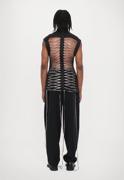 Vêtement sans manches noir avec un dos transparent et des détails de laçage argentés, complété par un pantalon large noir et des chaussures minimalistes.