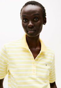 Camiseta polo amarilla con rayas blancas, de manga corta y con cuello. Presenta un pequeño logo en el pecho y una tela texturizada.