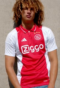 Maillot de sport rouge et blanc avec col polo, arborant un logo central "Ziggo" et l'emblème de l'Ajax avec trois étoiles au-dessus. Manches courtes.