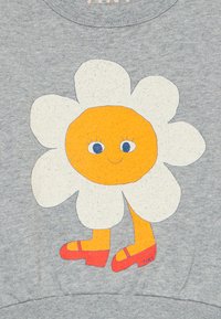 Weiße Gänseblümchen mit einem orangefarbenen lächelnden Gesicht, blauen Augen, gelben Beinen und roten Schuhen auf einem grauen Stoffhintergrund.