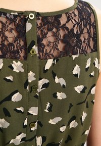 Camisa abotonada con un patrón de camuflaje en verde y beige. El panel de encaje negro en la parte posterior presenta detalles florales. Tejido suave con escote redondo.