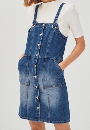 Kobieta ubrana w niebieską sukienkę na guziki z denimu z dużymi przednimi kieszeniami, na białym topie z krótkim rękawem i półgolfem.