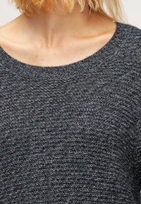 Mörkgrå stickad tröja med en strukturerad mönster, bred rund halsringning och ribbade muddarna. Nära bild av det övre plagget.