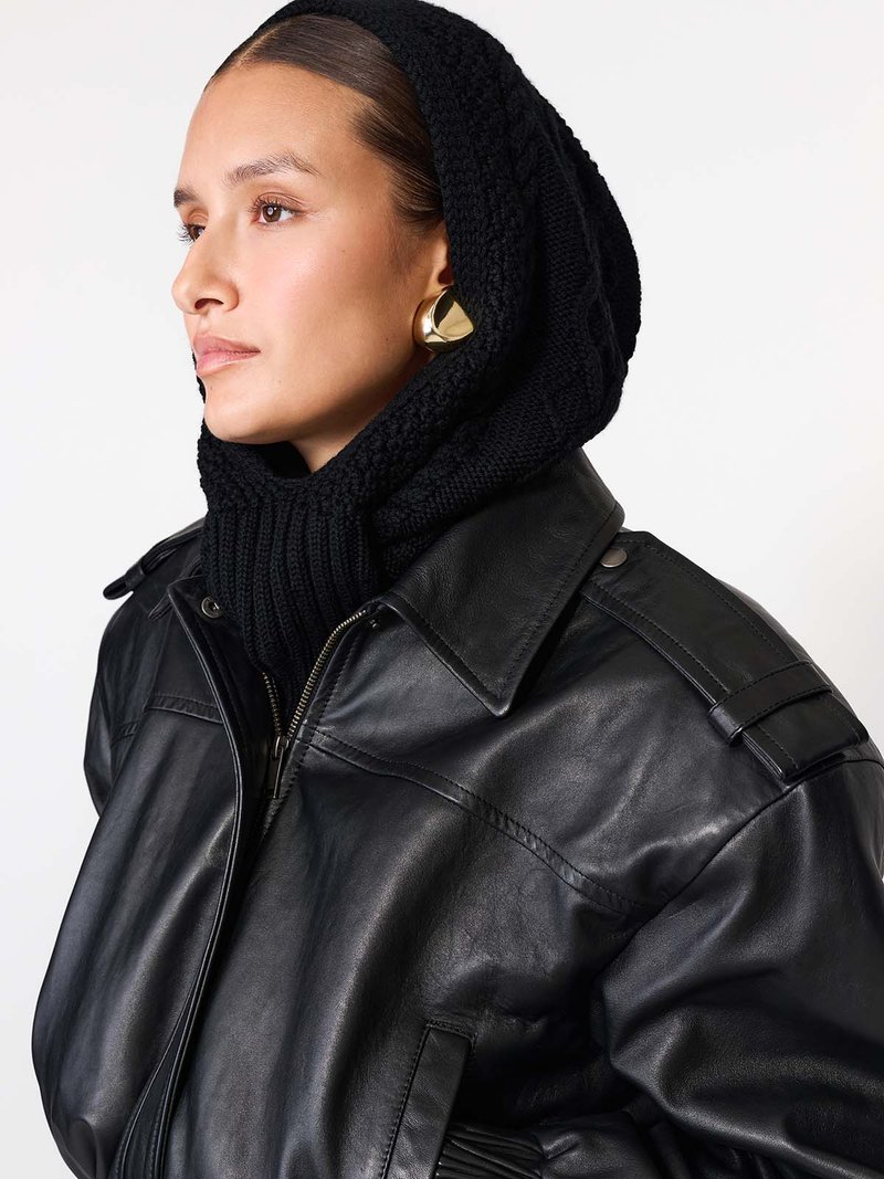 Fekete bőrdzseki strukturált gallérral és vállrészlettel, fekete kötött kapucnival és nagy arany fülbevalókkal párosítva.