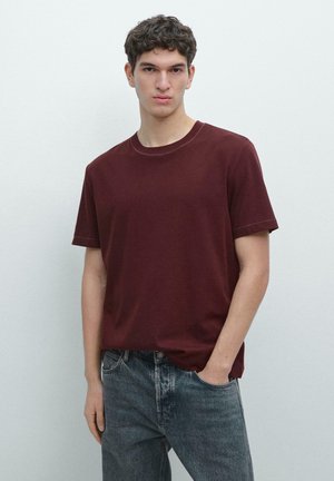 Jonge man met kort krullend haar, gekleed in een donkerrood T-shirt met korte mouwen en verwassen blauwe spijkerbroek, staand tegen een effen witte achtergrond.