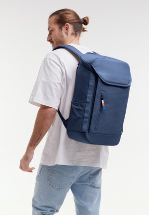 Man met een lichte huid en bruin haar in een knot, gekleed in een wit T-shirt en spijkerbroek, draagt een grote marineblauwe rugzak met meerdere vakken.