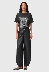 Mustassa graafisessa t-paidassa on leopardi-kuvio ja "ALLSAINTS"-teksti, yhdistettynä leveälahkeisiin mustiin nahkahousuihin ja tummiin liukusäädettäviin kenkiin, joissa on koristeellisia yksityiskohtia.