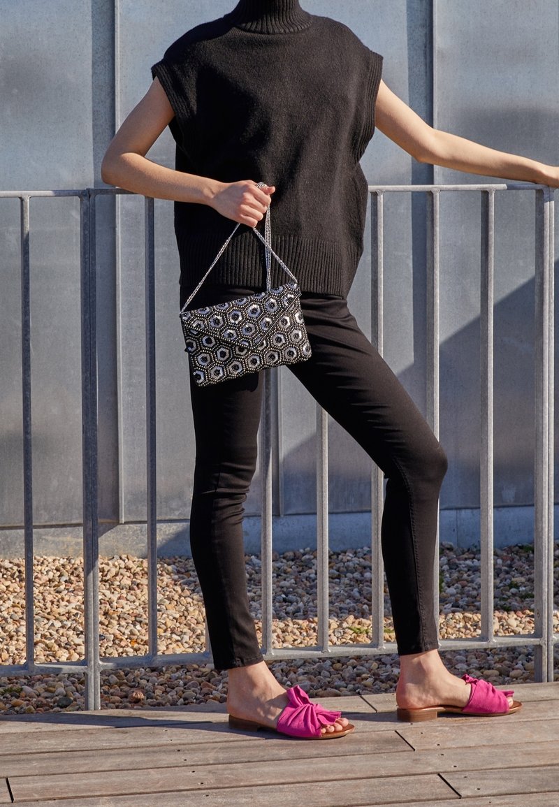 Femme debout près d'une rambarde en métal, portant un col roulé noir sans manches, un pantalon noir, des sandales roses et tenant un sac à main à motifs.