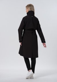 Zwarte trenchcoat met hoge kraag, een ceintuur in de taille en zichtbare stikdetails. Effen stof, knielang, gedragen over zwarte broeken en witte schoenen.