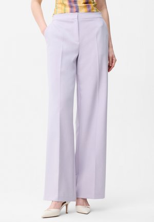 Kobieta ubrana w lawendowe spodnie z wysokim stanem i szerokimi nogawkami, połączone z kremowymi szpilkami w szpic oraz wielokolorową bezrękawną bluzką.