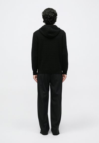 Sweatshirt preta com capuz, com padrão texturizado, apresentando um bolso frontal e capuz com cordão. Combinada com calças pretas largas.