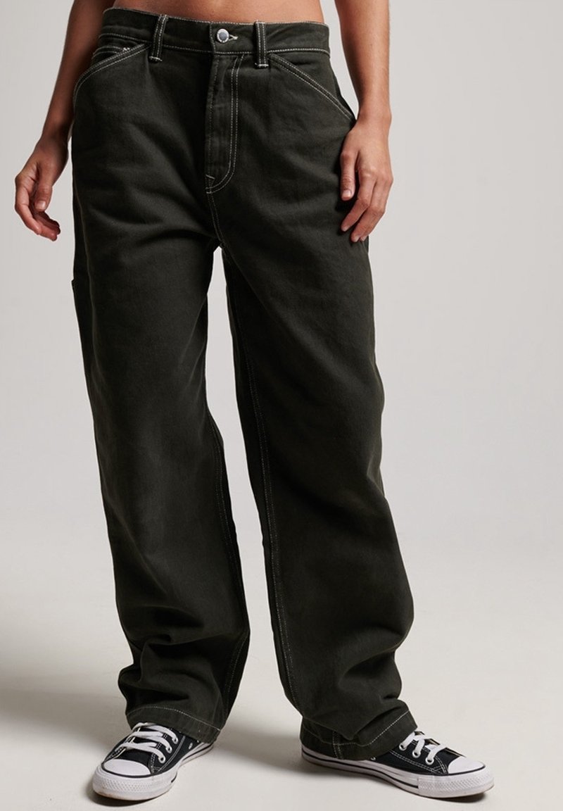 Personne portant un pantalon noir ample avec des coutures visibles et des baskets en toile noires à lacets blancs, se tenant devant un fond simple.
