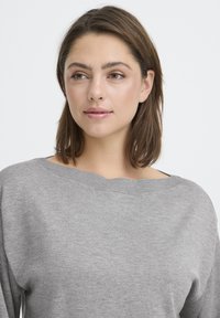 Noor naine, kellel on õlgadeni ulatuvad pruunid juuksed, kannab heledat halli kootud kampsunit ning vaatab veidi kõrvale valge tausta ees.