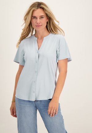 Vrouw met lang blond haar draagt een lichtblauw overhemd met korte mouwen en lichtblauwe jeans, staand tegen een effen lichte achtergrond.
