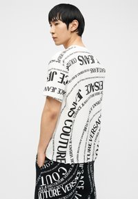 Νέος άνδρας με μαύρα μαλλιά, ντυμένος με λευκή και μαύρη στολή καλυμμένη με έντονα, ποικίλα προσανατολισμένα κείμενα «Versace Jeans Couture», στέκεται πλάγια.