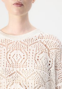Pull en tricot beige clair avec un motif en dentelle ouverte, un col côtelé et des motifs géométriques sur l'ensemble du tissu.