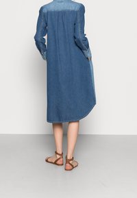Sukienka koszulowa z denimu z długimi rękawami, w jasnoniebieskim i ciemnoniebieskim gradientzie, z zaokrąglonym dołem i guzikami przy mankietach. Zestawiona z brązowymi sandałami na obcasie.