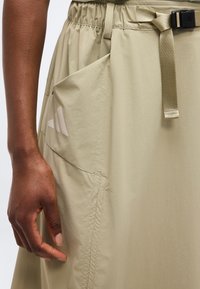 Κοντινό πλάνο μιας μπεζ φούστας με ελαστική μέση, ζώνη με ρυθμιζόμενο αγκράφα, πλαϊνή τσέπη και το χέρι ενός σκούρου χρώματος δέρματος που ακουμπά δίπλα της.