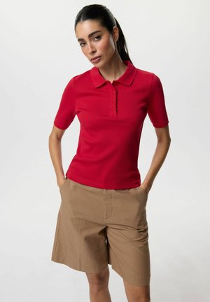 Kobieta z ciemnymi włosami związanymi w kucyk, ubrana w czerwoną koszulkę polo z krótkim rękawem i beżowe spodenki do kolan, stoi z rękami w kieszeniach.
