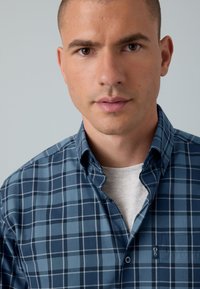 Blaues karierte Hemd mit knöpfbarem Kragen, das verschiedene Blautöne und weiße Linien aufweist, sowie ein kleines Logo auf der Brusttasche.