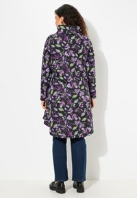 Abrigo de media longitud con estampado floral en negro, con acentos en púrpura y verde, y un cuello alto y mangas largas. Combinado con jeans oscuros.