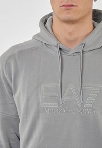 Felpa grigia con cappuccio caratterizzata da un logo ricamato, lacci con estremità in metallo e dettagli a coste lungo i polsini e l'orlo. Realizzata in tessuto morbido.