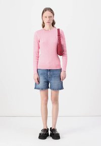 Rosa ullsweater i kabelstrikk, denimshorts og svarte platåkjoler. Modellen holder en rød crossover-bag. Enkel, moderne estetikk.