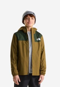 Αγόρι με καφέ και πράσινο μπουφάν North Face, γκρι σκούφο και μαύρο παντελόνι, στέκεται μπροστά σε λευκό φόντο.