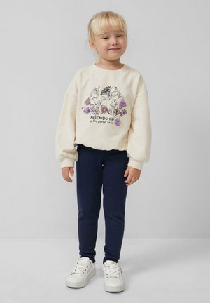 Felpa crema con stampa grafica che presenta tre personaggi, abbinata a leggings blu scuro e sneakers bianche. Tessuto morbido e vestibilità rilassata.
