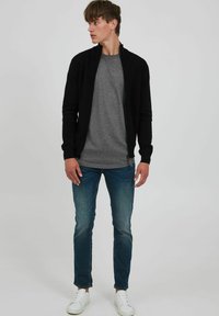 Chaqueta negra con cremallera y patrón texturizado, superpuesta sobre una camiseta gris. Combinada con jeans azules y zapatillas blancas. Silueta de corte ajustado.