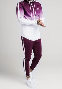 Henkilö, jolla on päällään violettiin ja valkoiseen liukuväriin oleva vetoketjullinen huppari sekä vastaavat housut, joissa on valkoiset raidat, seisoo kätensä edessä ristissä.