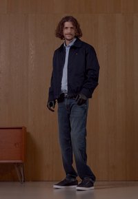 Marinejakke over en lys blå skjorte, mørke jeans og svarte sneakers. Glatt tekstur, enkel design og subtile merkevaredetaljer.