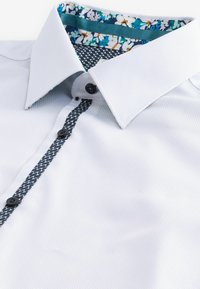 Weißes, strukturiertes Hemd mit einem gemusterten Kragen, der blaue und florale Designs aufweist. Die Vorderseite hat schwarze Knöpfe und einen kontrastierenden Besatz.