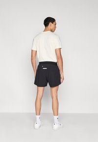 Μαύρο σορτς με λεία υφή και ελαστική μέση, συνδυασμένο με ανοιχτό μπεζ t-shirt· λευκές κάλτσες και αθλητικά παπούτσια ολοκληρώνουν την εμφάνιση.